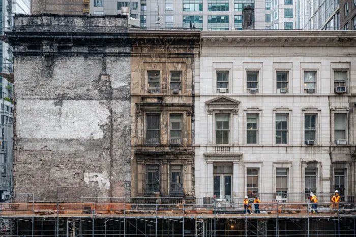 کارگران در حال مرمت و بازسازی نمای فرسوده ساختمان قدیمی در میان برج‌های مدرن شهری — پروژه ترمیم نما توسط گروه ساختمانی ساخَط