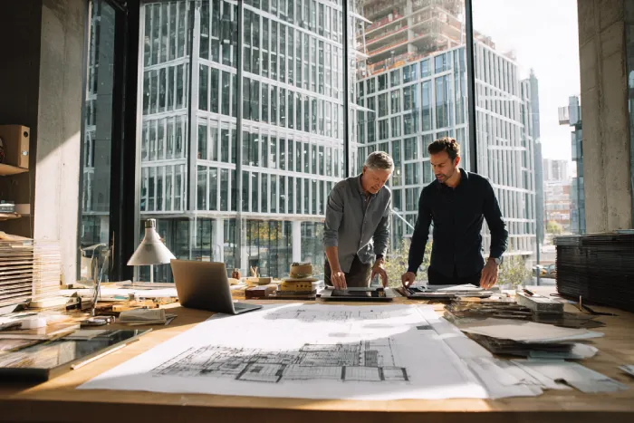 دو معمار در حال بررسی نقشه‌های فنی و جزئیات طراحی نما در دفتر معماری مدرن با چشم‌انداز برج‌های شهری — پروژه طراحی نما توسط گروه ساختمانی ساخَط